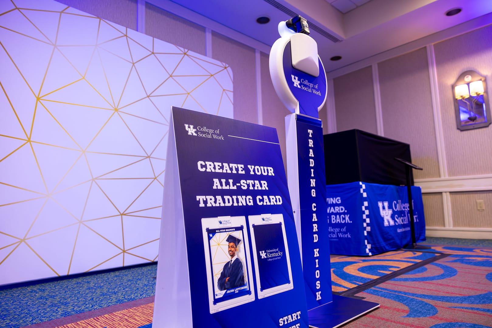 A trading card photo booth kiosk setup with a custom sign and custom table cover